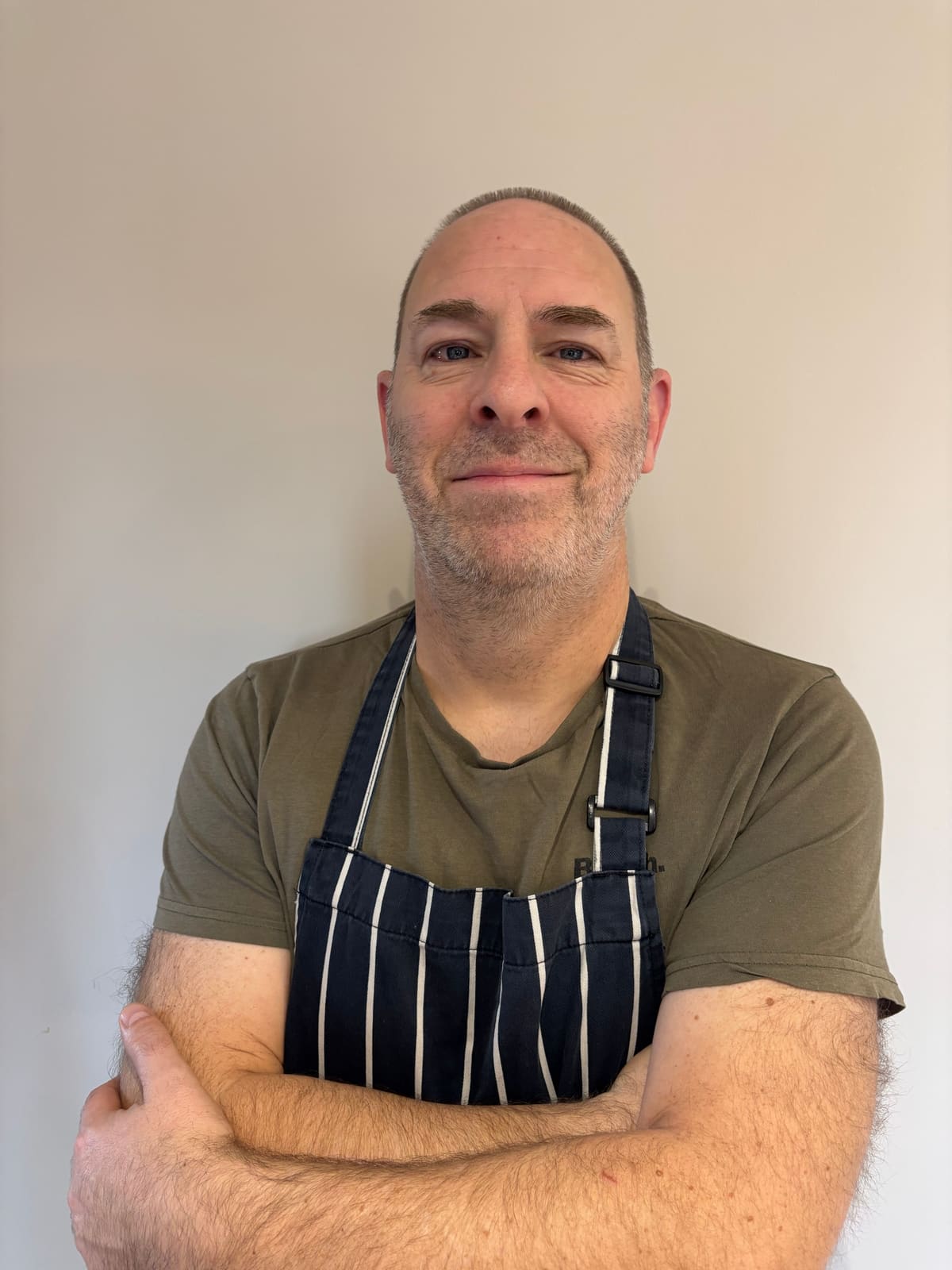 Chef Andrew Capper stands with folded arms wearing a striped apron.